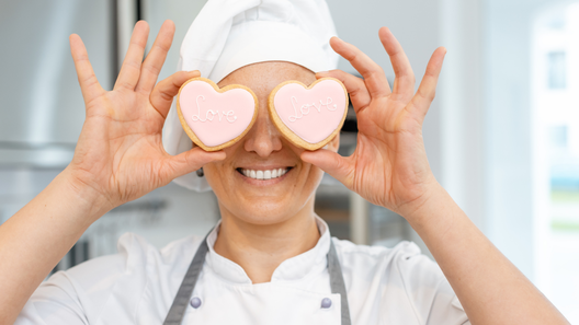 [alt=ハート型のアイシングクッキーを目にあてている笑顔のパティシエの姿。]

