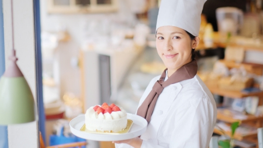[alt=ケーキを片手に笑顔でこちらを見ているパティシエの姿。]
